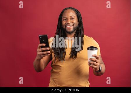 Photo of handsome cheerful smiling black man holding mobile phone in the hand and takeaway coffee tea cup, drinking hot beverage, in t-shirt, isolated over red background, chatting using front camera Stock Photo