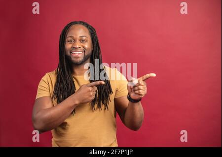Black man with beard dreadlocks looking at camera smiling with the index of both hands pointing to the side pointing to copy space in which to include your text, product or logo, concept of presenting Stock Photo
