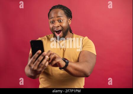 Amazed astonished dark skinned african man looking at the phone screen and typing, standing isolated over red background, wearing in casual mustard t-shirt, reading good pleasant unexpected message, Stock Photo