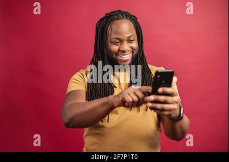 Happy dark skinned man looking at the phone screen, scrolling and typing message or information content, using smartphone for chatting, standing isolated over red background, search in the internet Stock Photo