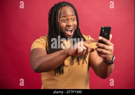 Excited smiling dark skinned man with long dreadlocks holding phone, reading good news in message, looking at screen, celebrating online lottery win, scrolling the information, on red background Stock Photo