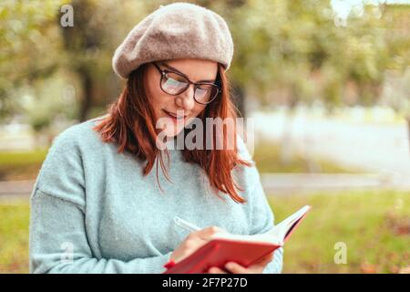 Portrait of young cute student girl writing notes in notebook sitting outdoors in nature park Stock Photo