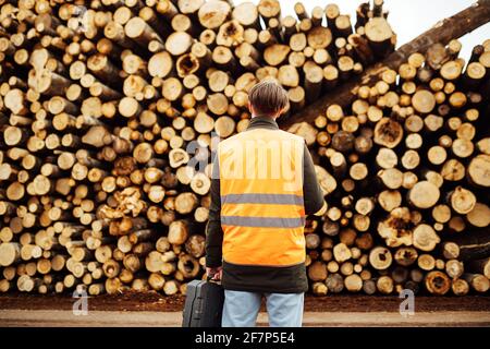 a worker in overalls stands with his back to the wood warehouse. a lot of logs harvested for woodworking at the factory. the man at the factory in rob Stock Photo