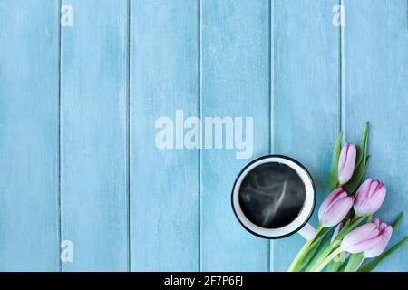 Steaming hot cup of coffee and pink spring tulip flowers over a rustic blue turquoise wooden table shot from top view. Stock Photo