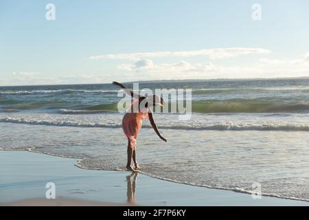 Mixed race woman wearing straw hat walking along the seashore with spread hands Stock Photo