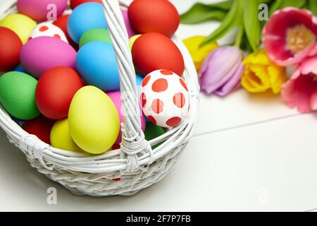 Easter eggs in basket and spring colorful flowers on white wooden table. Painted chicken eggs and multicolored tulips, Easter Day decor, selective foc Stock Photo