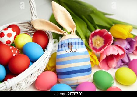 Easter bunny doll and Easter eggs in basket and spring colorful flowers on white wooden table. Painted chicken eggs and multicolored tulips, Easter Da Stock Photo