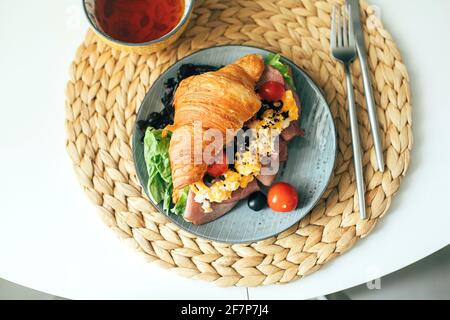 Fresh croissant sandwich with ham, scramble egg, tomato with tea on table, top view. Stock Photo