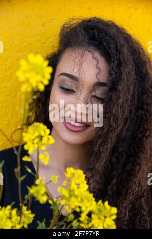 Beautiful woman with long curly hair standing behind delicate yellow blooming flowers Stock Photo