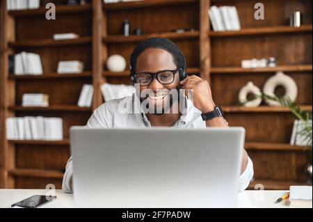 Happy African-American businessman in eyeglasses and headset talking via video call on the laptop sitting in the modern office, a friendly male entrepreneur connects with customers or colleagues Stock Photo