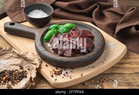 Slices of beef jerky, pepper, salt, and fresh basil on a beige chopping board. Jerky meat in a wooden plate. The concept of natural preservation of pr