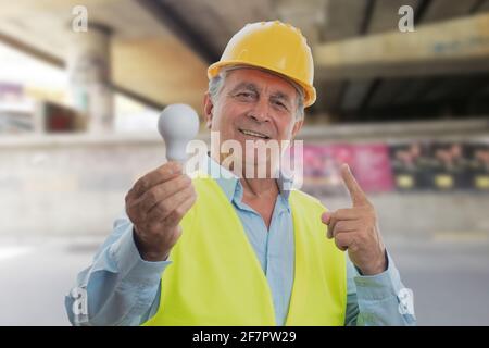 Happy old male constructor wearing yellow hardhat and vest holding lightbulb making good idea gesture using index finger Stock Photo