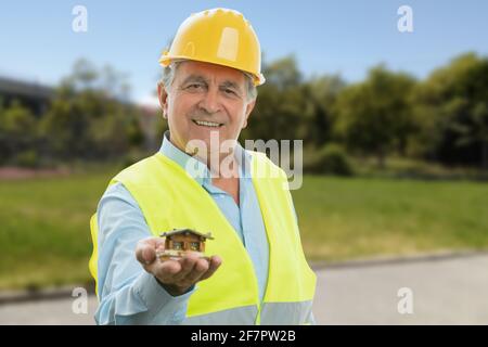 Old male builder in work attire smiling presenting miniature wooden house building concept park nature background Stock Photo
