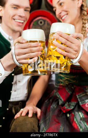 Young couple, man and woman, in traditional Bavarian Tracht drinking beer in a brewery in front of beer barrels Stock Photo