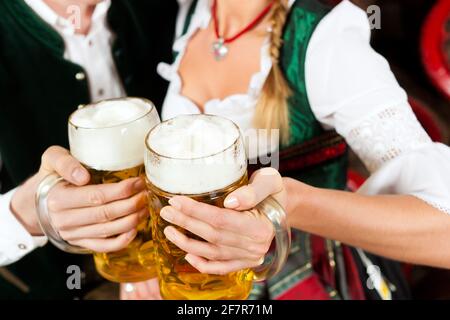 Young couple, man and woman, in traditional Bavarian Tracht drinking beer in a brewery in front of beer barrels Stock Photo