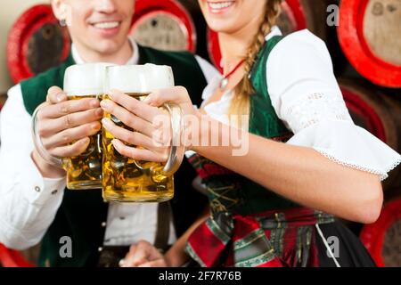 Young couple, man and woman, in traditional Bavarian Tracht drinking beer in a brewery in front of beer barrels Stock Photo