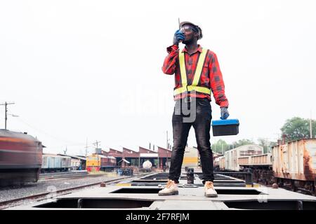 African machine engineer technician wearing a helmet, groves and safety vest is using a wrench to repair the train with talking by radio communication Stock Photo