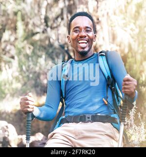 Portrait of a cheerfully laughing African-American Ethnicity young man sitting with a backpack and trekking poles and resting in the forest during a h Stock Photo