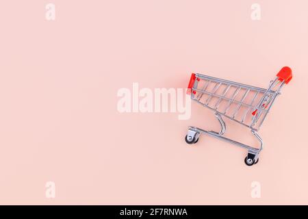 Shopping cart on beige background. Top view with copy space. Shop trolley at supermarket. Sale, discount, shopaholism concept. Consumer society trend. Stock Photo