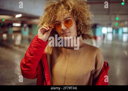Serious mixed race hip hop girl in jacket standing in garage and adjusting sunglasses while looking at camera. Stock Photo