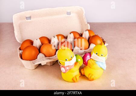 Conceptual Easter card. Two toy chickens in medical masks in quarantine with colored eggs on the table near the ecological packaging with orange chick Stock Photo
