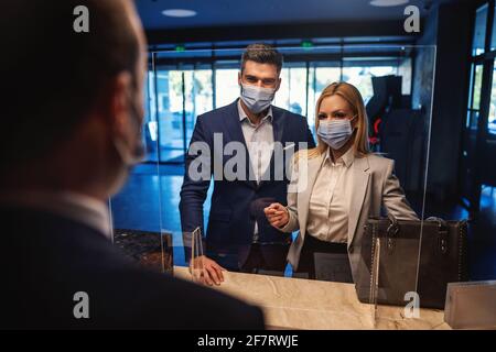 Business couple with face masks standing at reception and checking in the modern hotel. Business trip, relationship, travel during corona, COVID 19 si Stock Photo