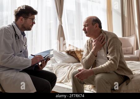 Doctor listening to elderly man complaints visiting him at home. Man suffering from throat pain. Stock Photo