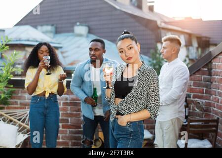 Charming young woman in stylish wear posing with glass of champagne on roof top. Three diverse friends drinking, dancing, chatting on background. Party celebration. Stock Photo