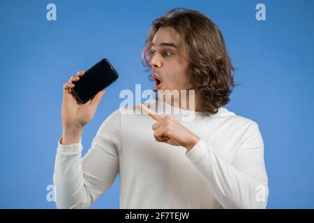 Handsome man pointing with finger on his mobile phone screen on blue studio wall. Modern technology, copy space. Stock Photo