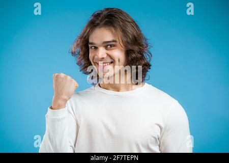 Man with long hairstyle is very glad and happy, he shows yes wow gesture of victory, guy achieved result, goals. Surprised excited happy guy on blue Stock Photo