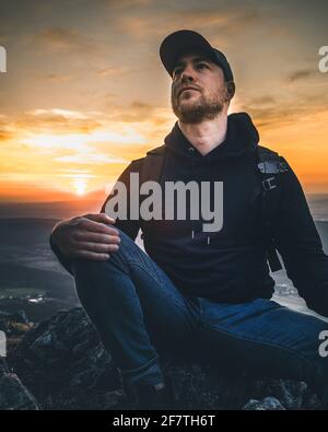 Man wearing a hoodie looking at a colorful sunset in mountains Stock Photo