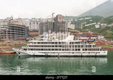 Yangtze River, Three Gorges, China - May 6, 2010: Xiling City. Wall of tall apartment buildings above shoreline with white ferry with bedrooms on gree Stock Photo