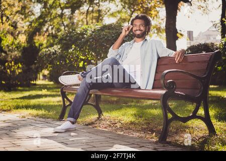 Portrait of attractive cheerful guy traveler sitting on bench touching specs sunny summer day outside outdoor Stock Photo