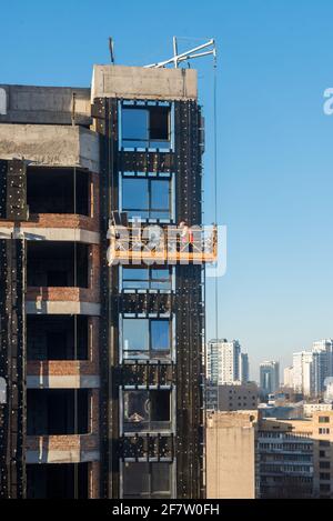 High rise construction work. Construction site workers in cradles working with facade. Suspended cradle for builders to work outside the skyscraper. B Stock Photo