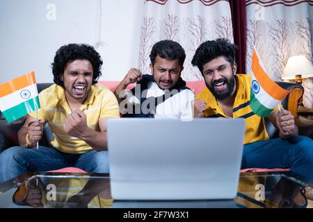 Three young people supporting Indian team by holding Indian flags while watching live streaming cricket match on laptop Stock Photo