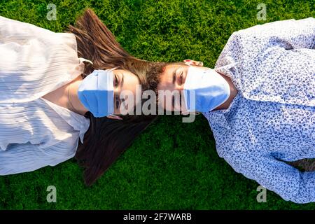 Beautiful couple of young students lying head to head on backs on green lawn of city park looking into the camera wearing the protective mask against Stock Photo