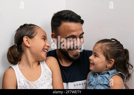 Caucasian father and daughters, looking at each other while smiling sitting on the floor and leaning against the white wall. Stock Photo