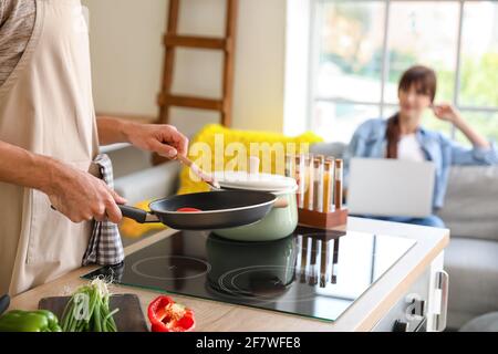 Young man cooking dinner while his wife is using laptop in kitchen Stock Photo