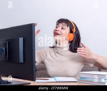 Attractive woman in orange headphones sitting at a table, looking at a computer monitor, gesturing, chatting on the Internet. Online adult learning co Stock Photo