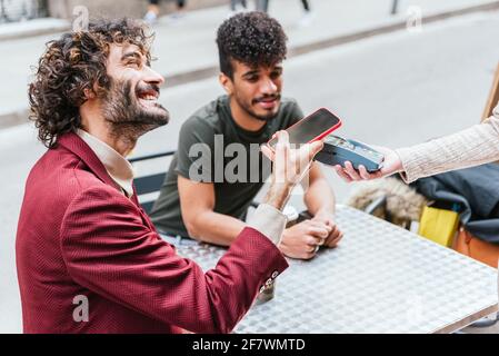 detail of a younc caucasian man using his phone to make a payment in a cafeteria. He is sitting in a terrace with a latin friend and smiles to unrecognizable waitress. Stock Photo