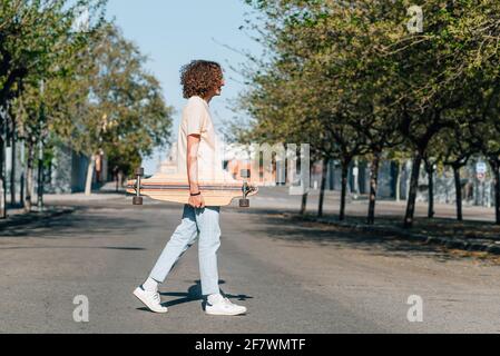 horizontal portrait of a red haired young man crossing the street. He has long curly hair. He is holding an skateboard Stock Photo