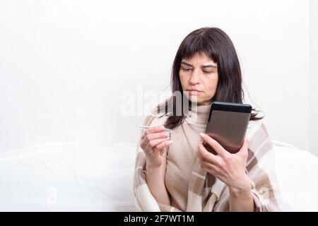 Portrait of a sick woman covered with a blanket holding a thermometer and communicating with the doctor on the tablet. Distance medicine concept. New Stock Photo