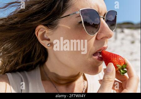 Close-up caucasian woman biting a strawberry. Girl in nature wearing sunglasses in a sunny, windy day. Stock Photo