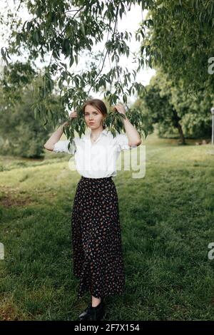 A young girl in a long skirt and a white shirt stands and holds with her hands to the branches of a tree and looks at the camera Stock Photo