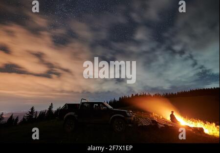 Wide angle view on cloudy starry night in the mountains and a man enjoying nature, sitting on logs in front of fire, off road car behind him. Concept of astrophotography, traveling and extreme ride Stock Photo