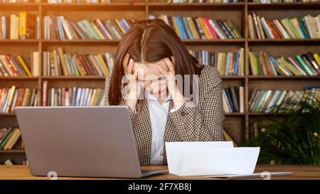 Businesswoman types answer on grey laptop looking at bank letter of account and puts head on hands in despair in office with racks of books Stock Photo