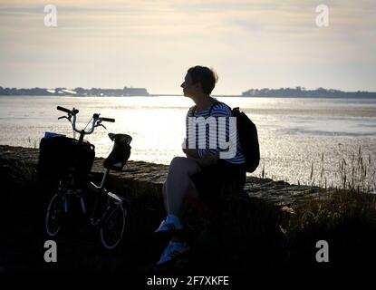 Frankreich, Batz-sur-Mer, 15.07.2019: Touristin mit Faltrad von Brompton auf einer Mauer im spaeten Gegenlicht am Rande des Marais Salants zwischen Ba Stock Photo