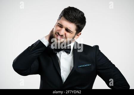 Tired young man having pain - neck ache. Guy in black holiday tuxedo takes break and kneads sore neck muscles with hand massage. Stock Photo