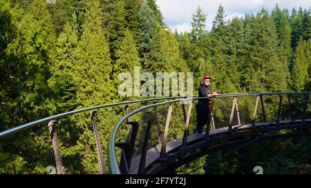 A young asian man in red hat and black clothes standing on a sky bridge, surrounded by tall green trees in Capilano Suspension Bridge Park of Canada. Stock Photo