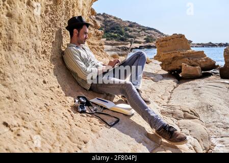A young journalist relaxes while telecommuting from the beach, beside him he has a camera, a notepad and a smartphone. Stock Photo